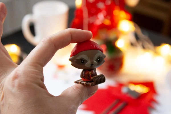 No Natal. Uma mão de close-up mantém uma figura de um brinquedo de guaxinim em um gorro vermelho em um trenó. Mesa de bokeh Gerlanda antes do jantar de gala — Fotografia de Stock