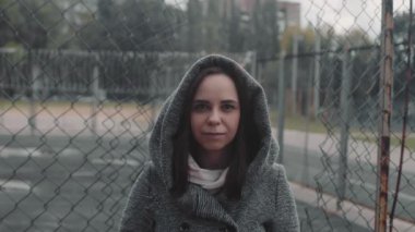 Kapüşonlu genç bir kadın kafes deliğinden kameraya bakıyor. Mantolu güzel bayan çitin yanında duruyor..
