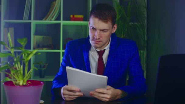 Joven examina documentos, sentado en la oficina. Hombre de negocios serio comprueba los papeles, sentado frente a la computadora portátil en el lugar de trabajo moderno. — Vídeos de Stock