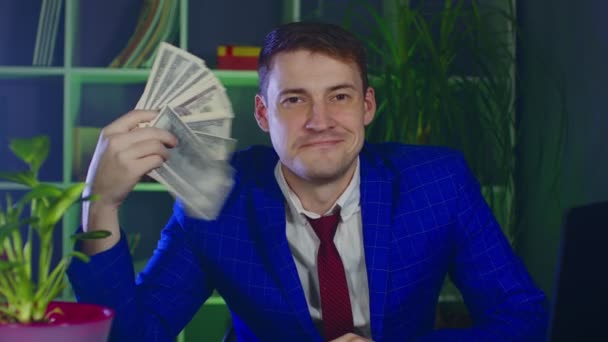 Homme d'affaires avec de l'argent regardant la caméra et souriant dans un lieu de travail moderne. Jeune homme salue l'argent en tant que fan, assis dans le bureau. — Video