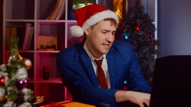 Junger Mann mit Weihnachtsmütze arbeitet am Laptop und tanzt, sitzt im Büro. Fröhlicher Geschäftsmann an modernem Arbeitsplatz in Neujahrsatmosphäre. — Stockvideo