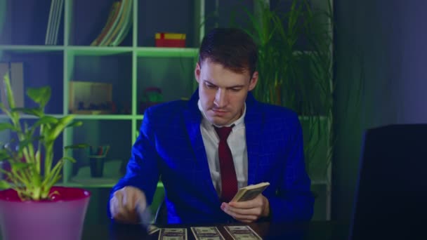 Genç iş adamı parayı sayıyor, ofisteki masanın üstüne yığıyor. Erkek girişimci dolar banknotlarını böler, modern işyerindeki masada oturur.. — Stok video