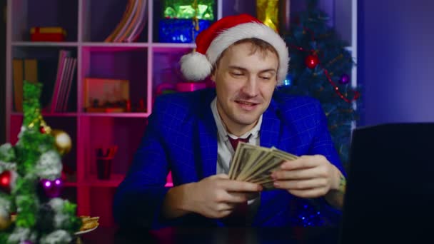 オフィスでお金とサンタの帽子の幸せなビジネスマン。クリスマスの帽子の男性起業家は、現代の職場でテーブルに座って、ドル札を保持します. — ストック動画