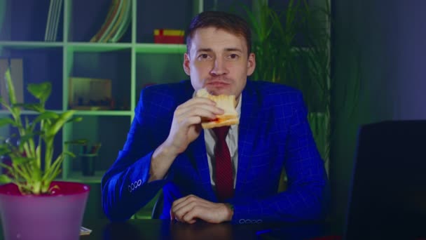 Молодий чоловік їсть сендвіч, сидячи в кабінеті. Бізнесмен насолоджується їжею на сучасному робочому місці під час роботи . — стокове відео