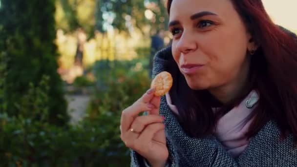 Potret seorang wanita cantik yang duduk di taman pada hari musim gugur yang cerah makan keripik kentang. Betina menempatkan keripik kentang di mulutnya pada latar belakang alam — Stok Video