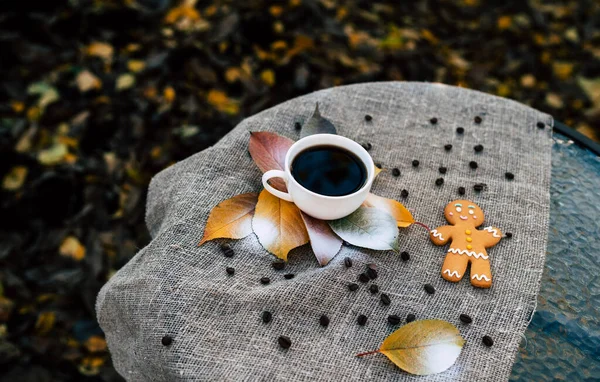 Пряничный человечек и чашка кофе возле осенних листьев. Вид сверху на сладкоежек и чашку крепкого кофе эспрессо, помещенные рядом с разноцветными осенними листьями на льняной ткани в парке — стоковое фото