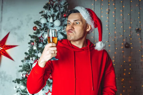 Jovem em Papai Noel com copo de champanhe no fundo da árvore de Natal. Homem adulto posando na árvore conífera com adornos decorativos. Conceito de celebração de Natal em casa. — Fotografia de Stock
