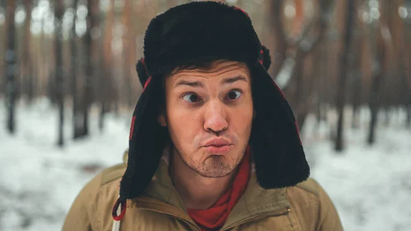 İnsan kış mevsiminde ormanda yüzünü buruşturur. Kulak kepi takan ve yüzünü buruşturan çılgın bir erkek portresi.. — Stok fotoğraf