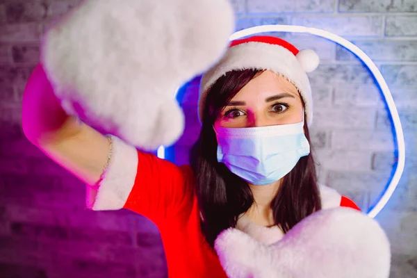 Femme en masque médical, costume du Père Noël montre signe de cadre contre mur illuminé.Femme en chapeau de Noël, masque de protection et gants. Concept de célébration de Noël en toute sécurité pendant la pandémie de coronavirus — Photo