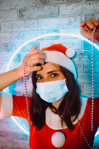 Donna in maschera medica e vestito di Babbo Natale con perline contro la parete illuminata.Primo piano della femmina in cappello di Natale e maschera protettiva.Concetto di festa di Natale sicura durante la pandemia di coronavirus — Foto Stock