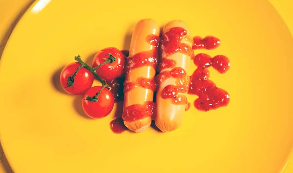 Primo piano di salsicce in salsa con pomodorini su piatto giallo. Frankfurters appetitosi con piccoli pomodori rossi e ketchup su fondo giallo. — Foto Stock