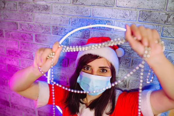 Женщина в медицинской маске и костюм Санта-Клауса с бусами против освещенных wall.Close женщины в рождественской шляпе и защитный mask.Concept безопасного празднования Рождества во время пандемии коронавируса — стоковое фото
