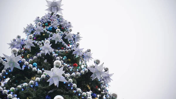 Árbol de Navidad con diferentes juguetes de Navidad contra el cielo nublado en el centro. Árbol de coníferas con adornos decorativos para crear ambiente festivo durante las vacaciones. —  Fotos de Stock