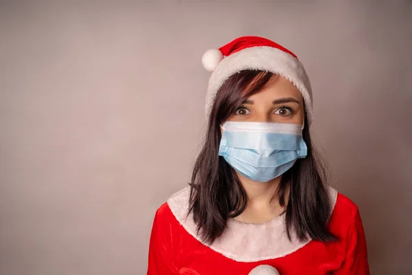 Молодая женщина в медицинской маске и костюме Санта Клауса на сером фоне. Крупный план женщины в Рождественской шляпе и защитной маске. Концепция безопасного празднования Рождества во время пандемии коронавируса. — стоковое фото