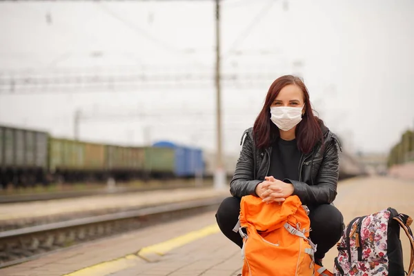 Mujer joven en cuclillas máscara médica en el andén, esperando tren. Pasajera en máscara protectora con mochilas sentadas en la plataforma del ferrocarril esperando el viaje en tren durante la pandemia de coronavirus —  Fotos de Stock