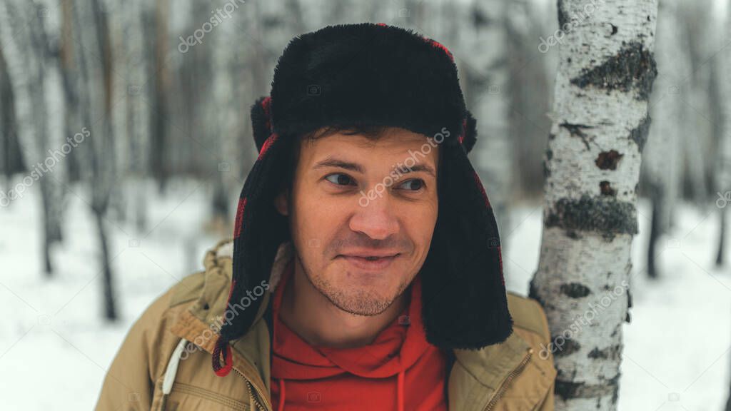 Man standing in forest in winter day. Serious young male wearing beige jacket over red hoodie and earflap hat standing on snowy forest and looking away