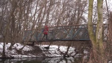 Genç bir kadın kış mevsiminde köprüden karşıya geçiyor. Kırmızı ceketli yetişkin bir kadın ağaçlık alanda yürüyor..