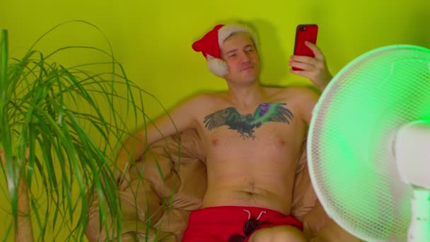 Ευτυχισμένος άνθρωπος με καπέλο santa με γυμνό κορμό παίρνει selfie στο smartphone, κάθεται στην πολυθρόνα πριν ανεμιστήρα. Χαλαρός άντρας φωτογραφίζοντας τον εαυτό τους στο κινητό τηλέφωνο και απολαμβάνοντας τις διακοπές του στο σπίτι, όπως στο θέρετρο — Αρχείο Βίντεο