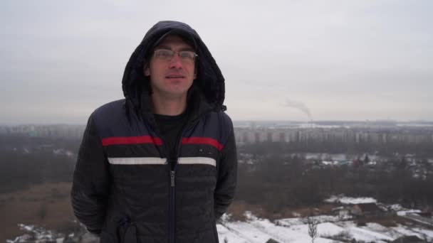 Mature man with glasses in hooded jacket looking thoughtfully into distance in cloudy weather. Adult male sniffing and thinking on background of panorama of small city. — Stock Video