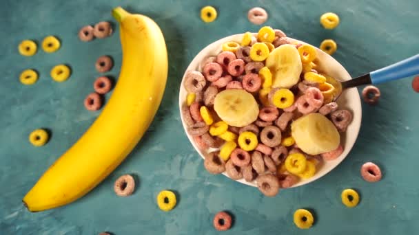 Здоровий сніданок з кашею та бананом. Зверху миска зі смачним здоровим сніданком, приготованим з різнокольоровими зерновими кільцями та бананом з молоком, поданим на синьому столі — стокове відео