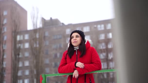 帽子と赤いジャケットを着た若い女性が通りに立っています。大人のブルネットは冬の季節に歩いて涼しい天気で暖まる. — ストック動画