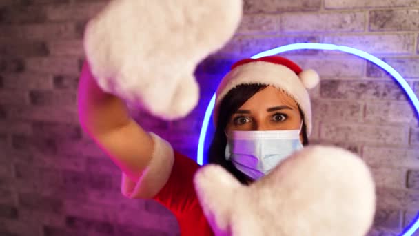 Tıbbi maskeli kadın, Noel Baba kostümü aydınlanmış duvara çerçeve işareti gösteriyor. Noel şapkalı, koruyucu maskeli ve eldivenli bir kadın. Koronavirüs salgını sırasında güvenli Noel kutlaması kavramı. — Stok video