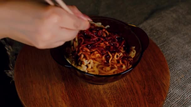 Fideos instantáneos listos con condimento y salsa picante en plato sobre tabla de madera. Mujeres irreconocibles mano con palillos baraja ramen con aditivos. De cerca.. — Vídeo de stock