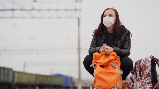 Ung kvinna i medicinsk mask knäböjer på perrongen, väntar på tåget. Kvinnlig passagerare i skyddsmask med ryggsäckar sittande på järnvägsplattform i väntan på tågresa under coronavirus pandemi — Stockvideo