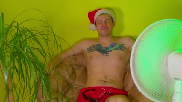 Щасливий чоловік у капелюсі Санта з голим торсом, сидить у кріслі перед вентилятором. Розслаблений чоловік із закритими очима насолоджується відпусткою вдома, як на курорті . — стокове відео