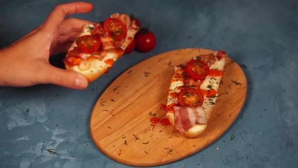 Životnost chutných bruschettů na dřevěné desce. Detailní záběr sendviče s roztaveným sýrem, slaninou, cherry rajčaty, koprem a omáčkou na řezací desce na modrém pozadí. — Stock video