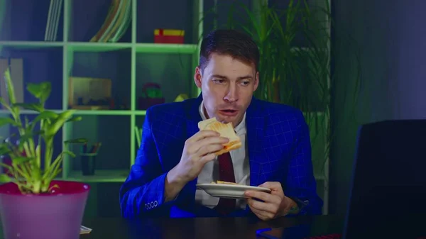 Young man eats sandwich, sitting in office. Businessman enjoying food at modern workplace during work. — Stock Photo, Image