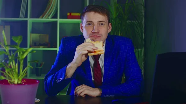 Jovem come sanduíche, sentado no escritório. Empresário desfrutando de comida no local de trabalho moderno durante o trabalho. — Fotografia de Stock