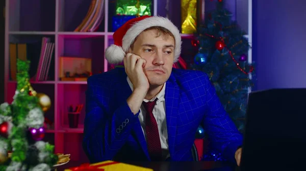 Noel Baba şapkalı genç adam dizüstü bilgisayarda çalışıyor, ofisinde oturuyor. Yeni yıl atmosferinde modern işyerindeki üzgün işadamı. — Stok fotoğraf