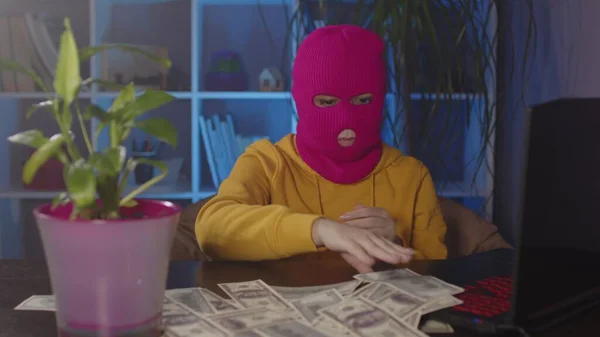 Jeune femme en cagoule rose recueille de l'argent de table au bureau. Pirate dans le masque avec des billets de dollars tout en étant assis dans un lieu de travail moderne. — Photo