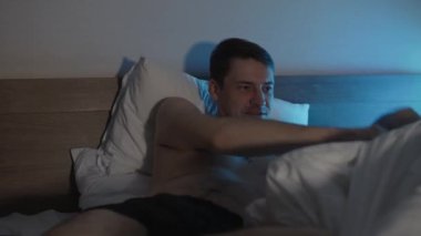 Genç adam yatakta yastığa uzanıyor, battaniye örtüyor ve akşam geç saatlerde televizyon seyrediyor. Rahatlamış erkek şovu karanlık odada izliyor..