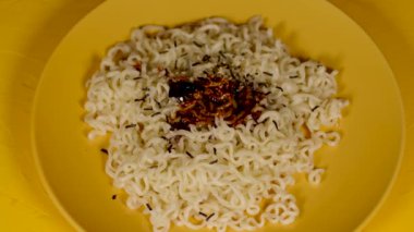 Sarı arka planda sarı tabakta baharatlı hazır şehriye. Tanınmayan biri baharatlı sosu tabağa ramen ile dolduruyor. Kapat..