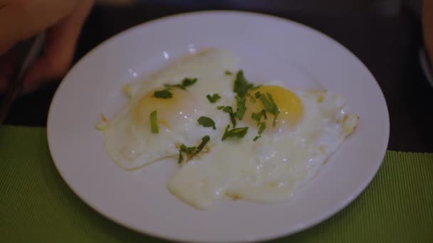 Fried eggs with parsley on white plate. Unrecognizable woman cuts off piece and eats. Breakfast concept. — Stock Video
