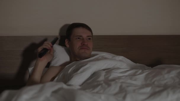 Young man lying on pillow under blanket in bed and watching tv in late evening. Relaxed male watching programs of show and switches channel with remote control in dark room. — Stock Video