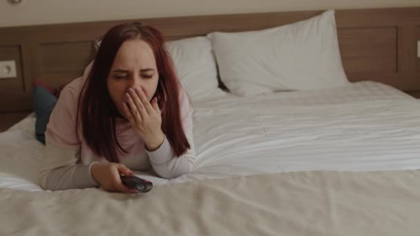 Young woman lying on bed, watching tv and yawning. Relaxed female yawns watching show, and switches channel with remote control. — Stock Video