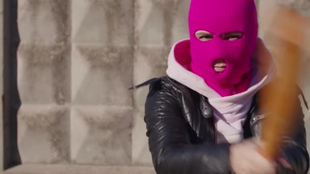 Молодая женщина в розовой балаклаве с бейсбольной битой в руках. Хулиган в маске бьется битой, смотрит в камеру. — стоковое видео