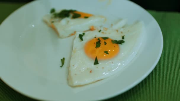 Fried eggs with parsley on white plate. Unrecognizable woman cuts off piece and eats. Breakfast concept. — Stock Video