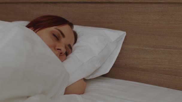 Una joven bajo una manta durmiendo en la cama por la mañana. Dormida hembra despierta y bosteza, acostada en la almohada. — Vídeos de Stock