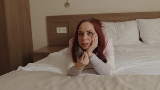Young woman in casual clothes lying on bed and watching tv. Emotional female watches favorite show, outraged and worried about film plot. — Stock Video