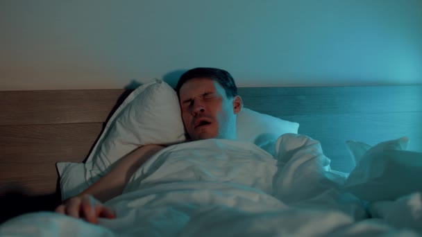 Jovem debaixo do cobertor a dormir na cama à noite. Homem sonolento boceja e acorda no quarto escuro. — Vídeo de Stock