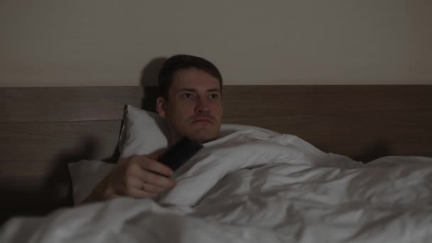 Ung mand ligger på pude under tæppe i sengen og ser tv i slutningen af aftenen. Afslappet mandlige se programmer for show og switches kanal med fjernbetjening i mørke rum. – Stock-video