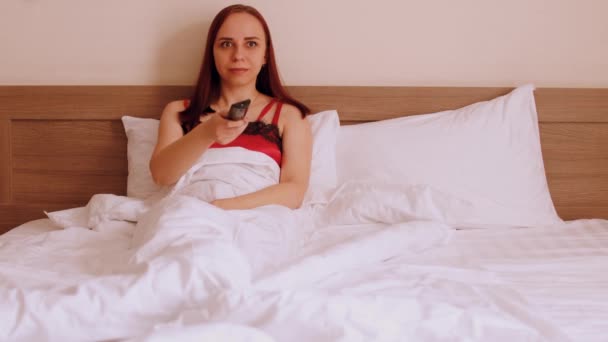 Genç bir kadın battaniyenin altında yatağa uzanıp televizyon izliyor. Rahat kadın izleme programları uzaktan kumandayla kanal değiştiriyor. — Stok video