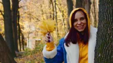 Sonbahar ormanında, ağacın yanında genç bir kadın. Gündelik giysiler içinde güzel bir kadın gülümser, sonbahar yapraklarını sallar ve fırlatır..