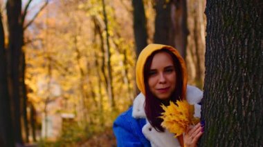 Sonbahar ormanında, ağacın yanında genç bir kadın. Güneşli ve rüzgarlı bir günde ağaç gövdesinin yanında dururken güler ve sonbahar yapraklarını tutar..