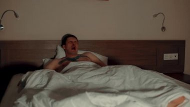 Yatakta battaniyenin altında yastığa uzanmış çıplak bir adam, gece geç saatlerde esniyor ve televizyon seyrediyor. Rahat erkek gözlerini ovuşturur ve televizyon izlerken uyumak ister..