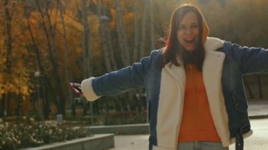 Genç bir kadın, şehir parkındaki kameraya bakıp gülüyor. Gündelik giysiler içinde güzel bir kadın güler ve sonbahar mevsiminde güzel havanın keyfini çıkarır..